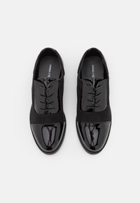 Chaussures noires avec une tige en daim et un capuchon en cuir verni brillant, lacées fermées et avec une semelle plate. La texture est lisse et mate.