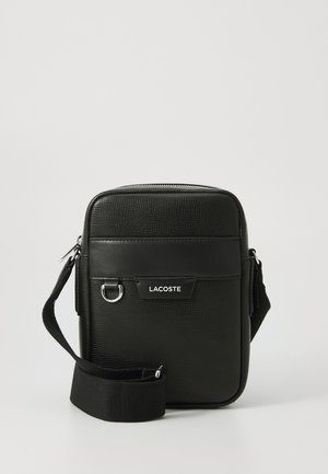 Geantă crossbody texturată, de culoare neagră, cu curea ajustabilă, buzunar frontal, detaliu cu inel metalic și logo-ul alb Lacoste pe panoul din față.