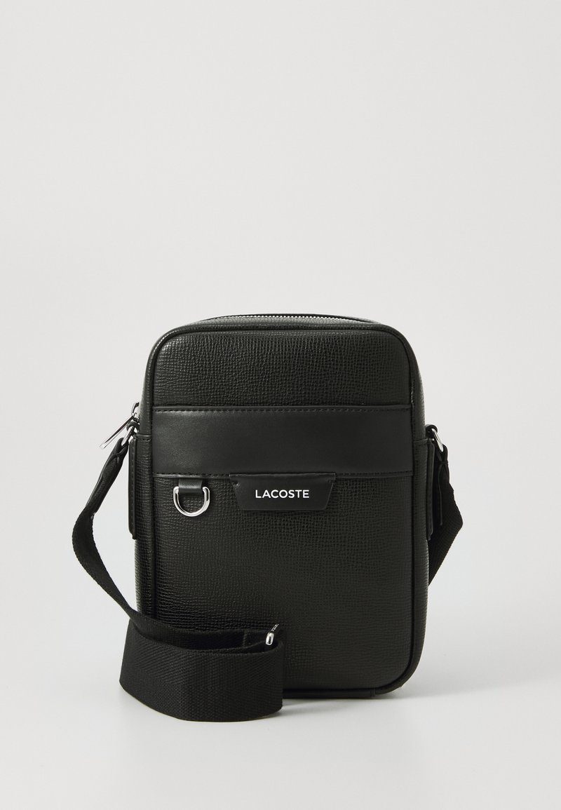 Sorte tekstureret crossbody taske med justerbar rem, frontlomme, metalringdetalje og hvid Lacoste-logo på frontpanelet.