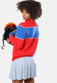 Maglione a righe rosso e blu con texture a costine, abbinato a una gonna blu plissettata con un motivo a quadretti. Casco da sci nero tenuto in mano.