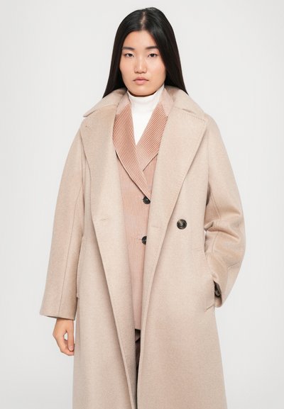 Beige wool blend coat with large lapels, double buttons, and side pockets, layered over a ribbed, light pink blazer.