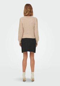 Donna con capelli di lunghezza spalle indossa un maglione lavorato a maglia beige, una gonna in denim nera e stivaletti neri con tacco alto, in piedi di spalle.