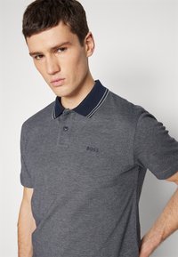 Polo krekls tumši pelēkā audumā ar tumši zilu apkakli, ar divu pogu kabatu un diskrētu logo kreisajā krūtī, ar teksturētu apdari.