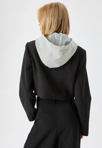 Blazer negro cortado con mangas largas y capucha gris. Presenta un ajuste entallado, textura suave y detalles de costura sutiles.