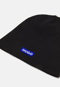 Beanie noire en tissu côtelé avec un patch rectangulaire bleu affichant le mot "HUGO" brodé. Design simple et texture douce.