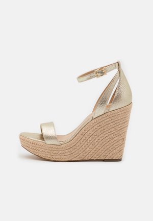 Guldmetallisk kilehæl sandal med flettet juteplatform, ankelrem og spænde lukning, vist fra siden på hvid baggrund.