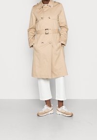 Trench coat beige con design doppiopetto, vita cintata e grandi bottoni, abbinato a pantaloni bianchi a 3/4 e sneakers beige.