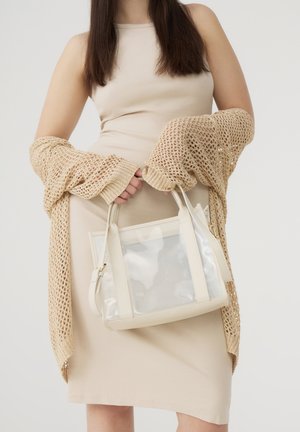 Borsa trasparente con dettagli crema, caratterizzata da una forma strutturata. Cardigan a maglia beige indossato sopra un vestito aderente beige.