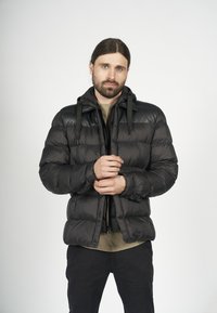 Svart pufferjacka med huva, med en glänsande läderövre del, framzipper och justerbara dragsnören, bärs över en beige skjorta.