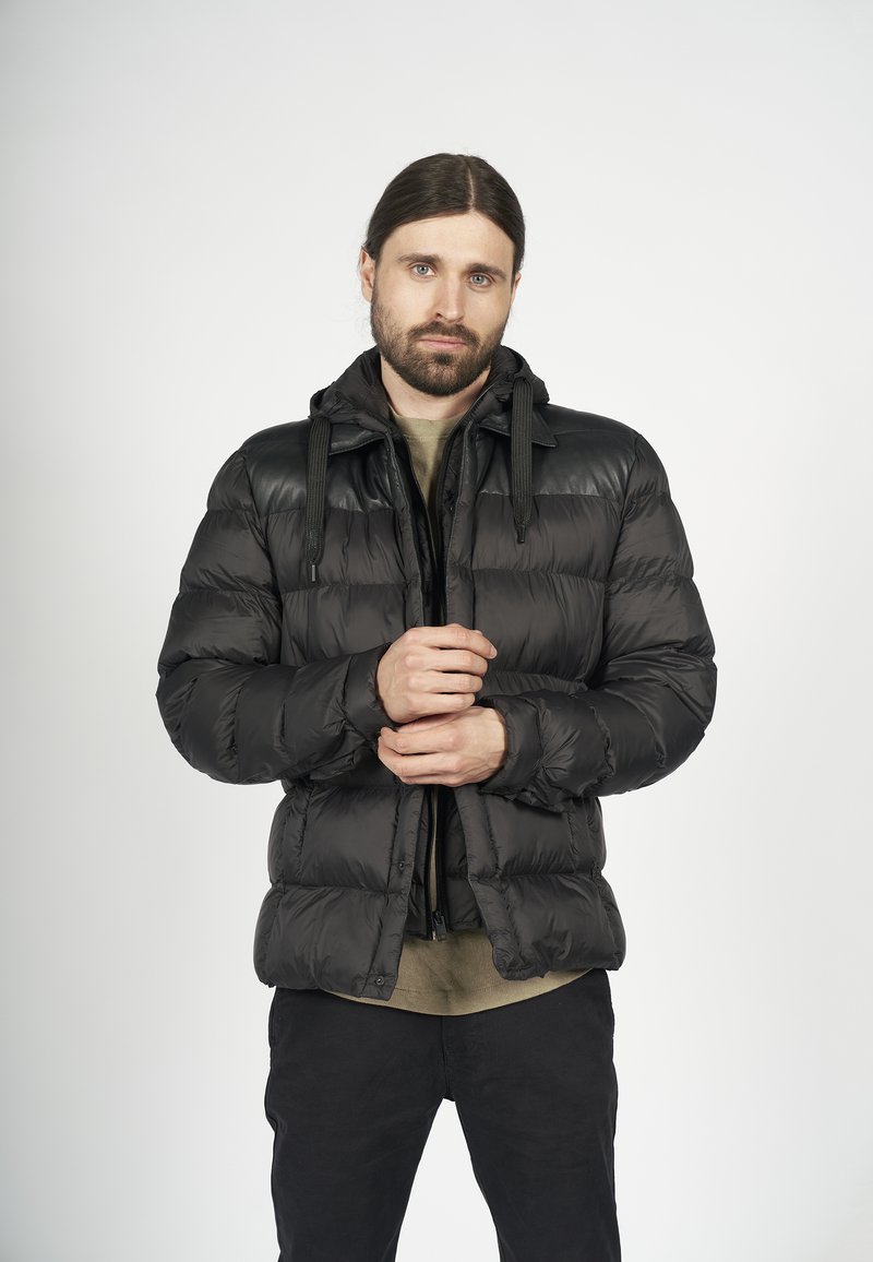 Svart pufferjacka med huva, med en glänsande läderövre del, framzipper och justerbara dragsnören, bärs över en beige skjorta.