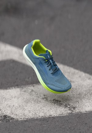 Blauer und neongrüner Sportschuh schwebt über einer Asphaltstraße mit weiß markierten Linien und wirbelt kleine Kieselpartikel auf.