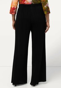 Pantaloni larghi neri con texture plissettata, vita elasticizzata e finitura del tessuto liscia. Vista da dietro, senza tasche visibili.