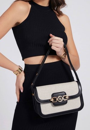Femme portant un crop top noir sans manches et une jupe, tenant un sac à main beige et noir avec une chaîne dorée et un fermoir logo.