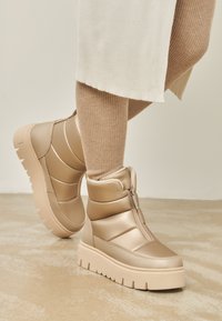 Botas de tobillo voluminosas beige con suela plana, cremallera lateral y paneles texturizados. La parte superior presenta un acabado suave y brillante.