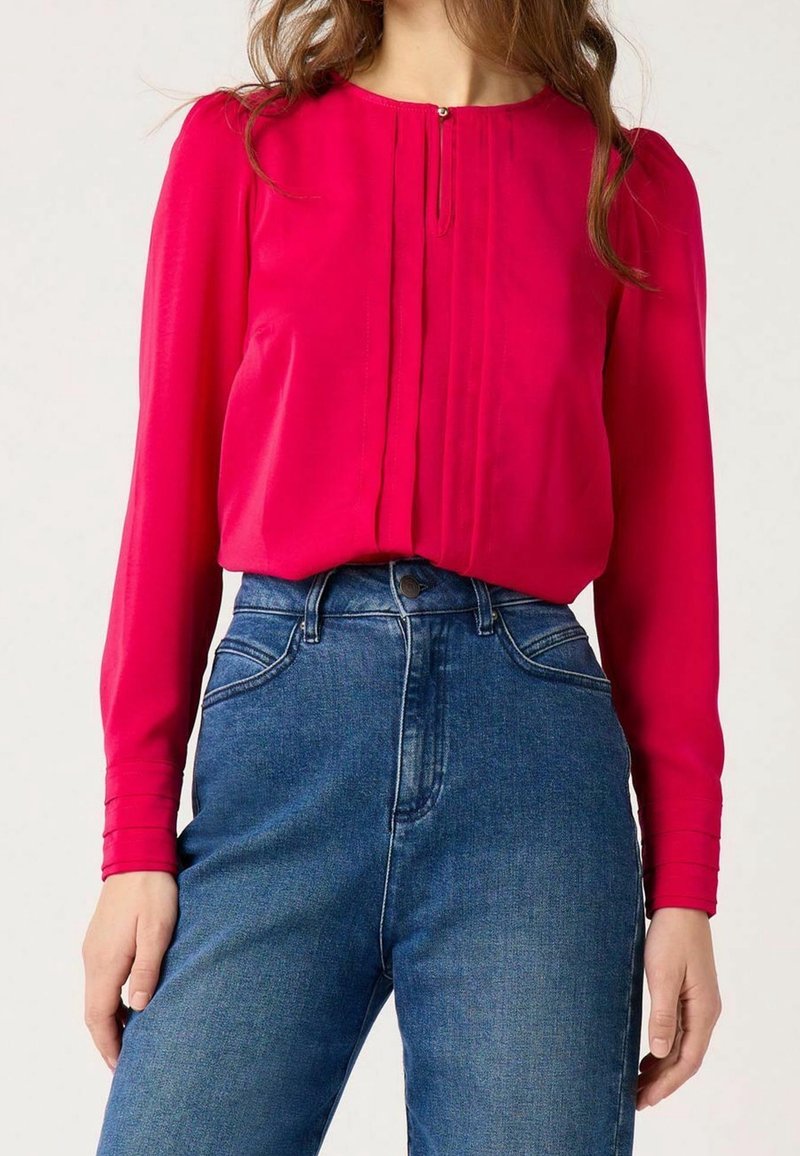 Bright pink long-sleeve blouse with pin-tuck detailing, round neckline, and fitted cuffs, paired with high-waisted blue denim jeans.