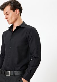 Camicia nera a maniche lunghe con bottoni, realizzata in un tessuto texturizzato, colletto all'italiana e bottoni abbinati. Indossata con una cintura nera.