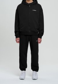 Zwarte zip-up hoodie met een kangoeroezak en logo op de borst, gecombineerd met bijpassende zwarte sweatpants. Gekozen met witte sportschoenen.