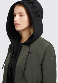 Maslinasto zelena bomber jakna s crnom kapuljačom. Sadrži zatvaranje na patentni zatvarač, crne vezice, izvezeni logo i rebraste naglaske.