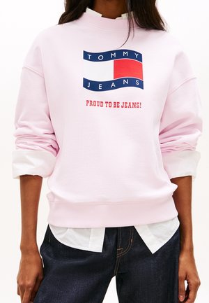 Osoba nosząca jasnoróżową bluzę z logo "Tommy Jeans" i tekstem "Proud to be Jeans!", w połączeniu z ciemnoniebieskimi dżinsami i białą koszulą.