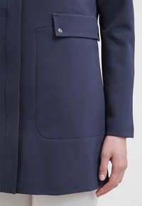 Tummansininen takki, jossa on läpällinen tasku ja metallinen nappi, jota kantaa henkilö, jolla on käsi kyljessä vakaassa taustassa.