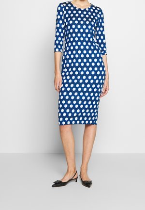Femme portant une robe cintrée mi-longue aux genoux avec un motif hexagonal bleu et blanc et des manches trois quarts, assortie à des chaussures slingback noires à talons bas.