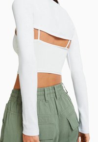 Kvinna i vit långärmad crop top över en vit linne, kombinerat med högmidjade gröna cargobyxor med fickor.