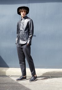 Chemise noire en cuir à boutons avec poches latérales, assortie à un pantalon sombre. Fini avec des chaussures noires à semelles et lacets blancs.