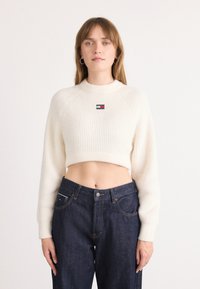 Cream ribbed knit crop sweater with a high neckline and raglan sleeves, featuring a small logo patch on the chest and paired with dark denim jeans.