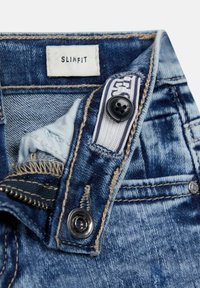 Dettaglio di jeans in denim blu slim-fit con zip, chiusura a bottone e etichetta bianca all'interno della vita.