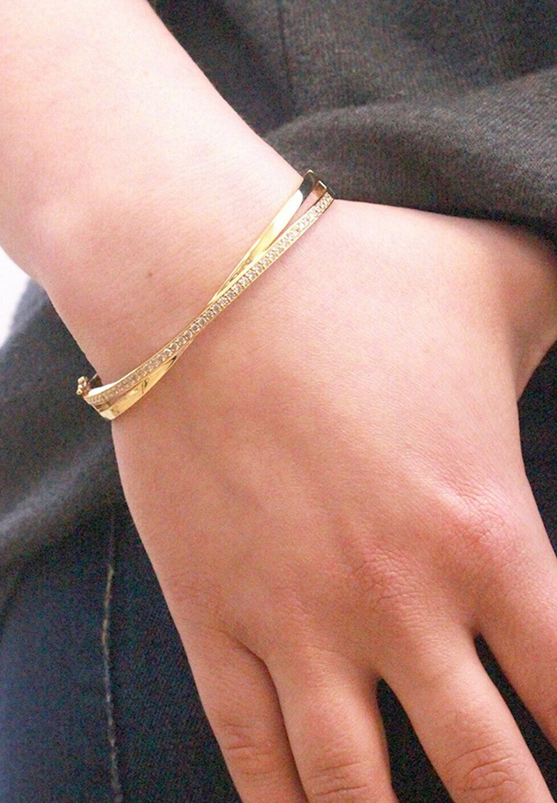 Gouden armband met een slanke, gedraaide vorm, voorzien van een rij kleine heldere edelstenen aan de ene kant. Glad metalen oppervlak, minimalistische esthetiek.