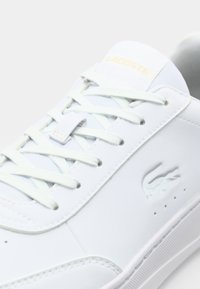 Valkoinen nahkainen tennari, jossa litteät nauhat, prässätty logo ja teksturoitu ulkopohja. Ominaisuuksina kullanvärinen teksti ja minimalistiset design-yksityiskohdat.