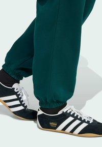 Sorte og hvide ruskinds sneakers med gummisål og tre hvide striber, parret med grønne joggers med snøre, der har elastiske manchetter.
