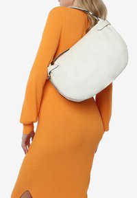 Bolso de mano de cuero blanco con forma curva, correa delgada y herrajes plateados, llevado sobre un vestido naranja de canalé.