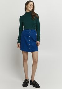 Grüner Rollkragenpullover kombiniert mit einem blauen Kordrock mit Knopf, der einen geraden Schnitt und Fronttaschen hat. Schwarze Loafers mit goldenen Akzenten.