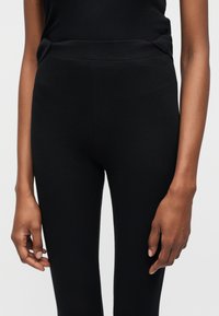 Γυναίκα που φοράει μαύρα κολάν με υψηλή μέση, στέκεται με τα χέρια χαλαρά στα πλευρά, μπροστά σε απλό φωτεινό φόντο.