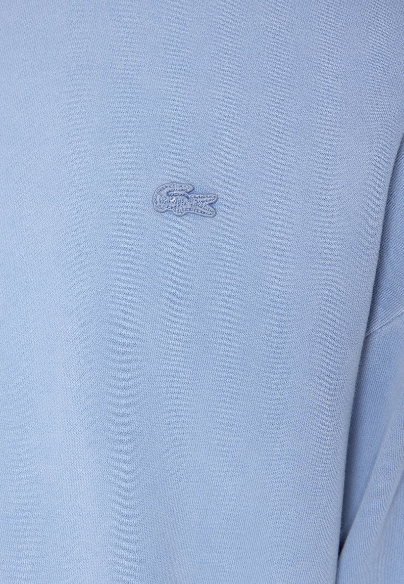 Tela azul clara con un pequeño logo de cocodrilo bordado en el área del pecho de una prenda.