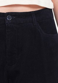 Gros plan d’une personne portant un pantalon en velours côtelé noir taille haute avec un bouton et une poche avant visibles sous un top blanc.