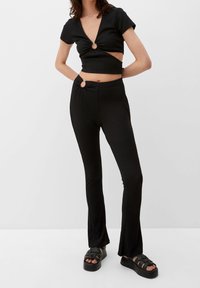 Černý dvoudílný outfit se skládá z přiléhavého topu s výstřihem do V, kruhovým výřezem a kalhot s vysokým pasem a rozšířenými nohavicemi. Hladká textura.