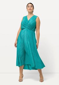 Vestido en tono verde azulado con detalle plisado, diseño de escote en V, sin mangas y cintura fruncida, que llega hasta la mitad de la pantorrilla, combinado con sandalias de tacón plateadas.