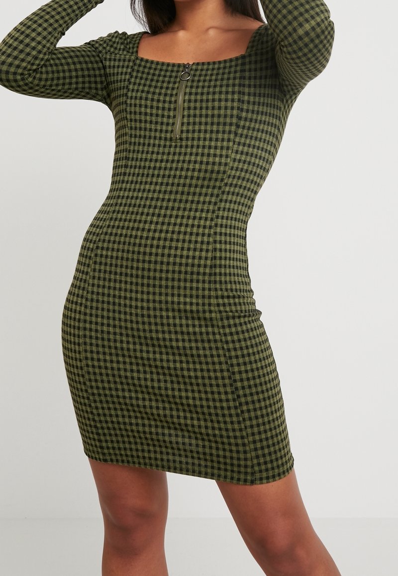 Robe à carreaux verts et noirs, design ajusté, manches longues, encolure carrée, avec une fermeture éclair à l'avant. Tissu texturé.