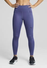 Puma vijolične legice z visokim pasom, gladkim materialom in kontrastnim črnim logotipom, v kombinaciji s črnimi športnimi čevlji.
