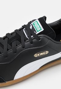 Sneaker in pelle nera con accenti bianchi, dotata di lacci neri piatti, un logo dorato "KING" e una suola in gomma con texture color gomma.