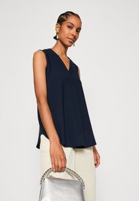 Haut sans manches bleu marine avec un décolleté en V et une coupe décontractée, associé à un pantalon de couleur claire et un sac à main en argent texturé.