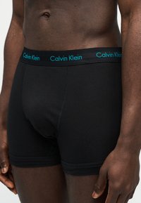 Ανδρικός κορμός με σκουρόχρωμο δέρμα που φοράει μαύρα μποξεράκια Calvin Klein με κορδέλα μέσης σε τιρκουάζ χρώμα με λογότυπο, στέκεται μπροστά σε απλό φόντο.