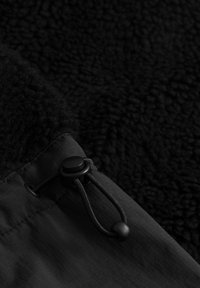 Tessuto felpato nero con finitura testurizzata, dotato di un toggles con cordino e bordi in materiale liscio e piatto, il tutto in una palette monocromatica.