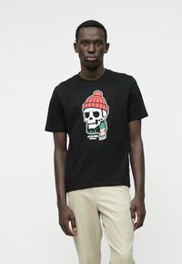 Čierne bavlnené tričko s grafikou lebky v červenej pletenej čiapke. Dizajn obsahuje farebné akcenty a text na spodnej časti.