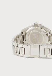 Stříbrné nerezové ocelové hodinky s náramkem na články, kulatým pouzdrem a texturovanou korunkou. Zadní strana je zdobena vyrytým logo a lesklým povrchem.