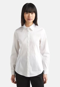 Camicia bianca a maniche lunghe con colletto classico, vestibilità dritta, chiusura frontale con bottoni e tessuto di cotone liscio. Indossata con pantaloni scuri.