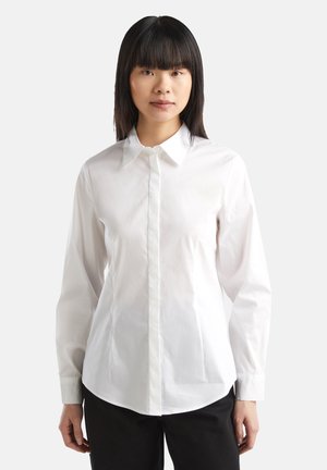 Chemise blanche à manches longues avec un col classique, coupe droite, fermeture à boutons sur le devant et texture en coton lisse. Portée avec un pantalon foncé.