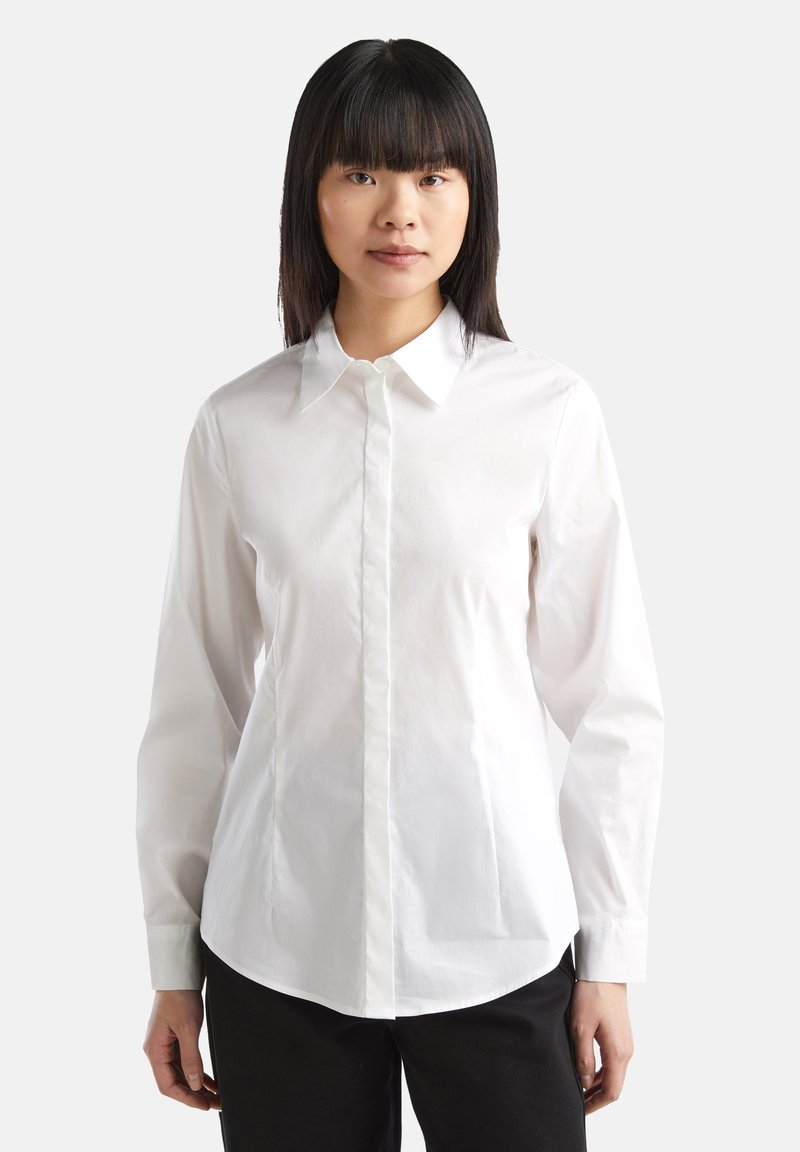 Camicia bianca a maniche lunghe con colletto classico, vestibilità dritta, chiusura frontale con bottoni e tessuto di cotone liscio. Indossata con pantaloni scuri.